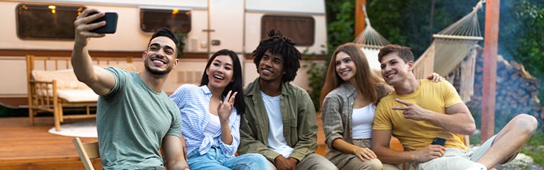 Diverse young friends taking selfie near campfire in evening, using smartphone, sitting next to RV during camping trip