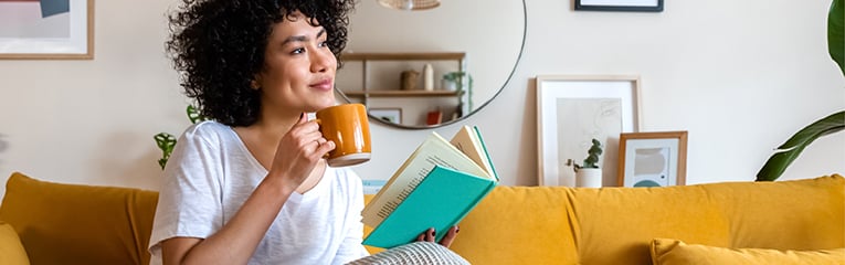 Pensive relaxed African american woman reading a book at home, drinking coffee sitting on the couch