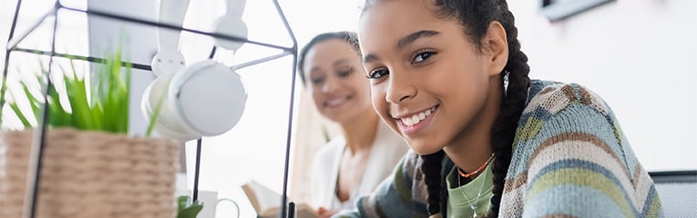 happy african american teen girl looking at camera writing in notebook near blurred mom with book